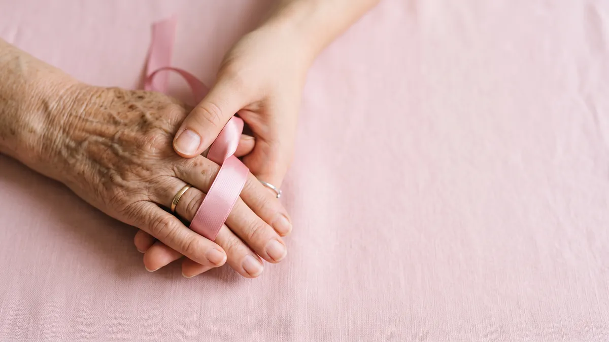 Mão jovem apoiada sobre uma mão idosa, com uma fita rosa entre os dedos